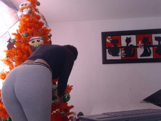 VanneSamChaud se inclina, vistiendo una camiseta negra y leggings grises, mostrando su trasero. Está decorando un árbol de Navidad naranja con adornos verdes. En la pared hay un cuadro con la silueta de un gato..