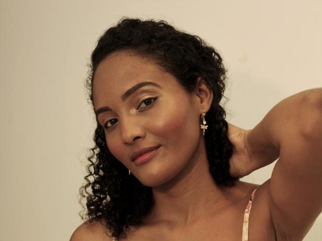Megan Gil tem cabelo preto encaracolado, pele morena e está sorrindo com a mão atrás da cabeça. Ela está usando brincos de cruz dourados e uma blusa rosa com alças..