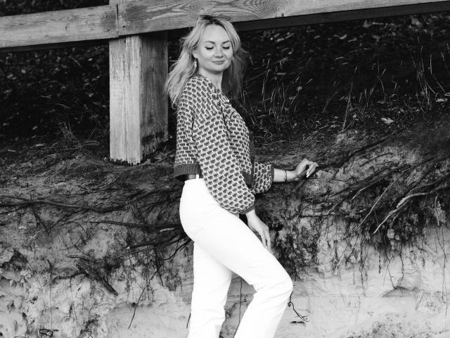 KayaBlonde, avec ses cheveux blonds, portant une blouse à motifs et un pantalon blanc, se tient près d'une clôture en bois, tenant un bâton, montrant ses fesses, regardant vers le bas avec un léger sourire..