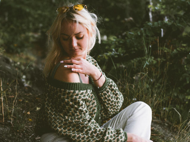 KayaBlonde with blonde hair, green sweater, and white pants, sits in forest, hand on chest, yellow sunglasses on head, looking down, soft natural light, calm expression.