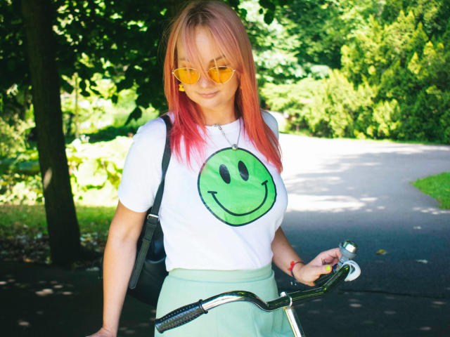 KayaBlonde with pink-dyed hair, yellow sunglasses, white tee with green smiley, green skirt, holding bike, black backpack, standing on sunny path, smiling slightly.