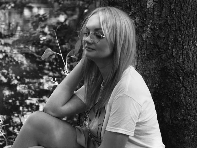 foto em preto e branco de Kaya: loira, óculos, camiseta branca, shorts, sentada encostada em uma árvore, segurando uma folha, relaxada, com um leve sorriso, mão no queixo, fundo de floresta, joelho levantado, casual..
