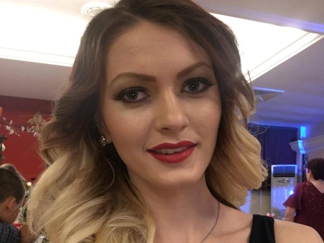 BlossomPussy hat blondes, welliges Haar, roten Lippenstift und schwarzen Eyeliner. Sie trägt ein schwarzes Oberteil und blickt mit einem leichten Lächeln in einem hell erleuchteten Raum in die Kamera..