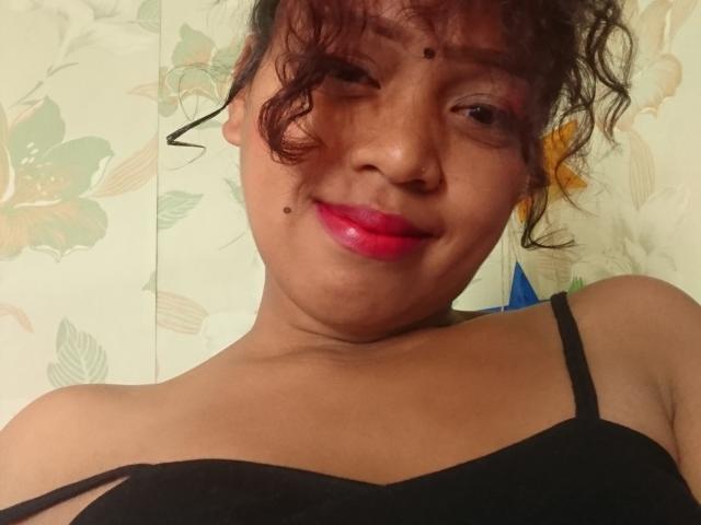 Mahaitia, avec ses cheveux bruns bouclés, son rouge à lèvres rouge, son haut noir à bretelles et ses petites taches de rousseur sur le visage, sourit à l'appareil photo, montrant un léger double menton..