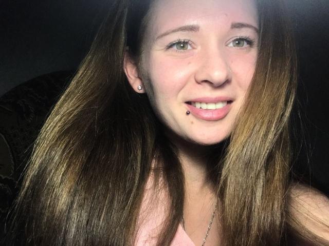 HereIsSoul has long brown hair, a nose piercing, and a labret piercing with a small bead. She's smiling with pink lips, green eyes, and wearing a pink top.