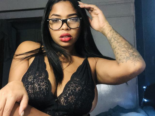 VickyGreys with black lace bra, red lipstick, glasses, and arm tattoo, touching her hair, showing her big tits. Dark-haired, medium skin, looking at camera. Bedroom setting.