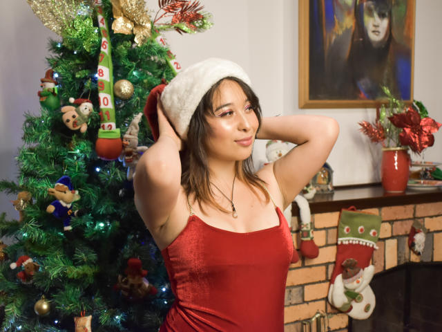 Chloe Lavigne, con i capelli castani, indossa un abito di velluto rosso e un cappello da Babbo Natale bianco, con le mani tra i capelli, in piedi di fronte a un albero di Natale decorato. Il suo seno è visibile..