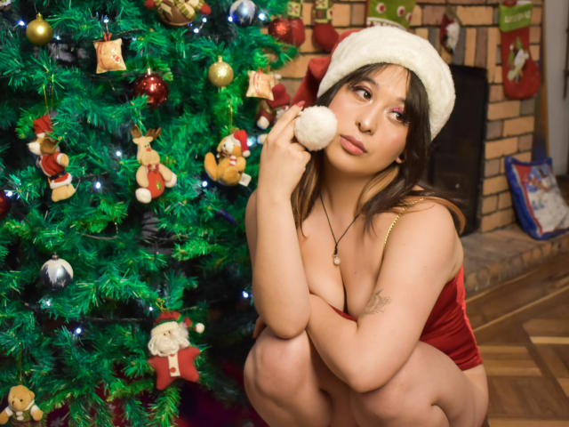 ChloeLavigne, in een hurk, draagt een rode fluwelen top, een witte hoed, houdt een pluizige pompom vast, kijkt naar de camera. Links staat een kerstboom met versieringen en lichtjes..