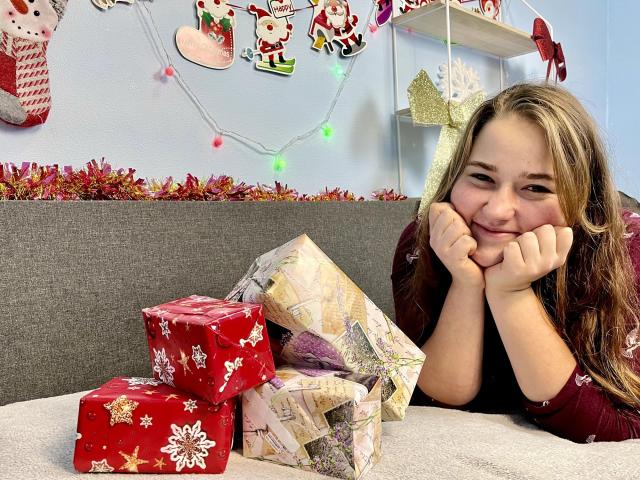 DaniellaGraceZZ with long brown hair, smiling with hands on cheeks, lying on a grey couch, looking at wrapped Christmas presents on a white table, wearing a maroon top.