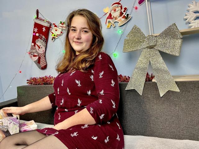 DaniellaGraceZZ, chubby with medium tits, wearing a red dress with white patterns, sitting on a gray couch, holding a gift, with Christmas decorations and a large silver bow.