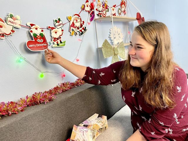DaniellaGraceZZ with brown hair, wearing a maroon top, pointing at Christmas wall decor, smiling, sitting on gray sofa, colorful string lights, glittery white ornament, and wrapped gift.