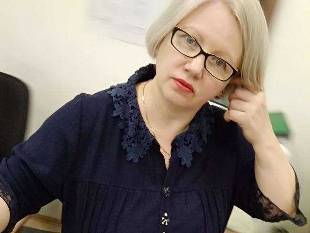 Agnnessa, com pele clara, cabelo loiro e óculos, vestindo uma camisa azul escura com cadarços, tocando sua orelha com a mão direita, batom vermelho e um leve franzir de lábios..
