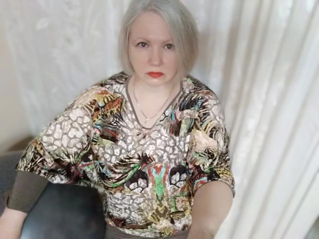 Agnnessa, met bleke huid en grijs haar, draagt een kleurrijke, gestreepte blouse, heeft rode lippenstift en een neutrale uitdrukking, en zit op een zwarte stoel..