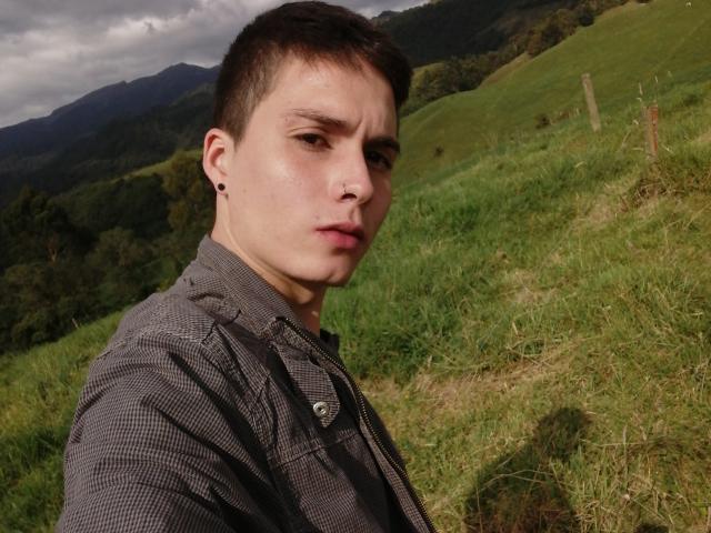 JackRouse, con cabello castaño corto, aretes negros, piercing en la nariz y expresión seria, vistiendo una camisa a cuadros, de pie en un campo cubierto de hierba con montañas al fondo..