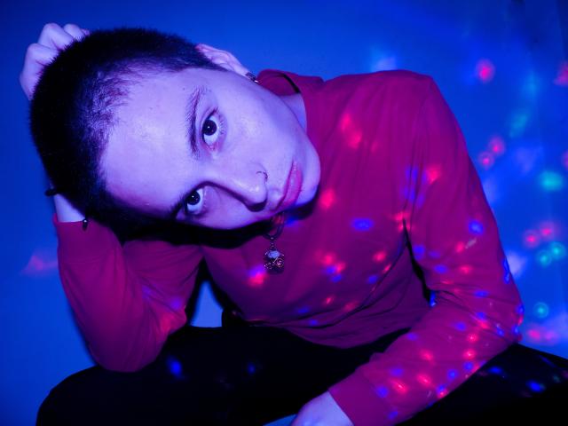 JackRouse, con cabello corto y oscuro, ojos azules y un piercing en la nariz, vistiendo una camisa roja de manga larga, sentado con la mano en la cabeza, rodeado de luces láser de colores..