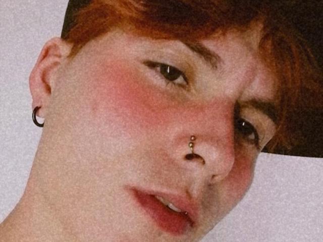 JackRouse tiene el pelo rojo, un piercing en la nariz y un pequeño aro en la oreja. Su rostro está enrojecido, con la boca ligeramente abierta y un pequeño piercing visible en el labio..
