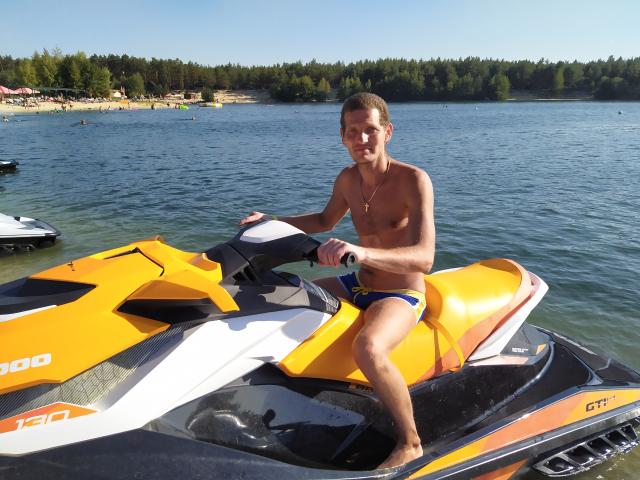 PierceBrick, zonder shirt, draagt een blauwe zwembroek, zit op een gele en zwarte jetski, houdt het stuur vast, er is water voor hem, een bos op de achtergrond, een heldere lucht, daglicht, kort bruin haar, een ketting..