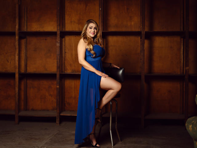 LylaGreen, com seios grandes, vestindo um vestido azul, sentada em uma cadeira, com uma perna levantada, sorrindo, com cabelo castanho ondulado, batom vermelho, saltos pretos, em um quarto de madeira..