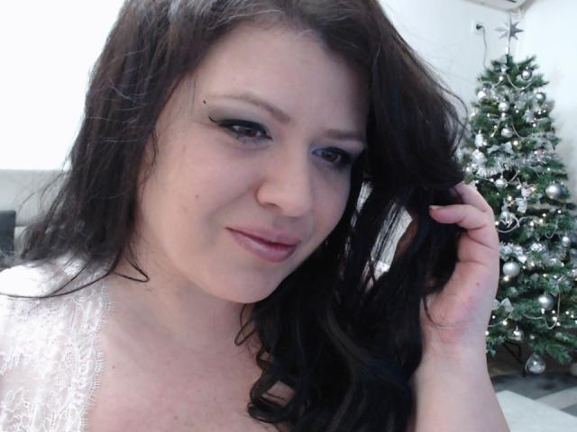 Carolina69 mit langem, schwarzem Haar, einem weißen Spitzenoberteil und schwarzem Eyeliner, leicht lächelnd, die Haare haltend, im Hintergrund ein Weihnachtsbaum mit silbernen Ornamenten..