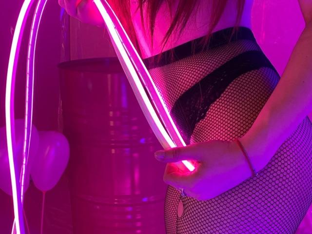 Alisa Mertes sosteniendo tiras de luz rosa neón contra su trasero cubierto con medias de red, vistiendo encaje negro, con iluminación rosa y púrpura, y su pulsera roja visible en su muñeca derecha..