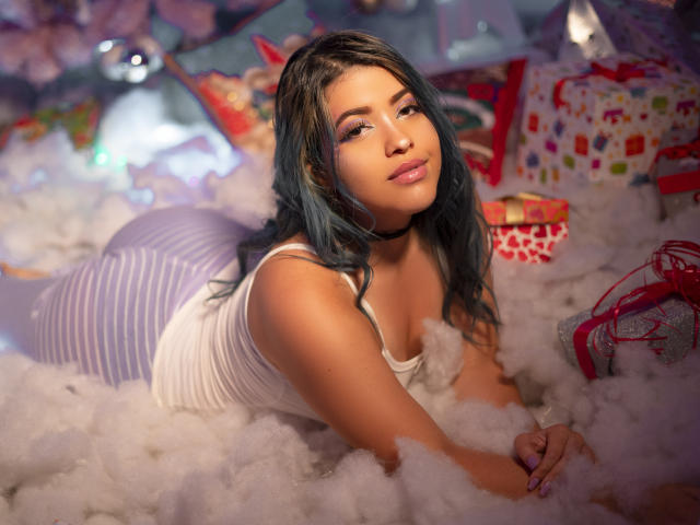 KimLafayette con i capelli blu, una canotta bianca e pantaloni a righe viola e bianchi, sdraiata in un "fumo" bianco soffice, con un'espressione sexy e sicura, labbra rosa chiaro e una collana nera..