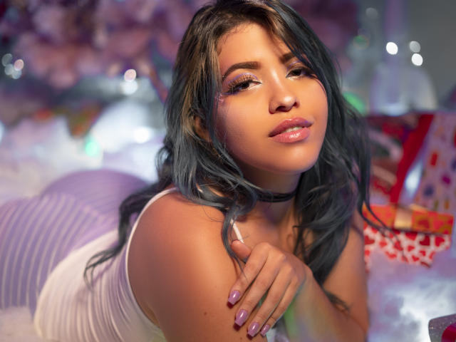 KimLafayette, avec ses longs cheveux bleus, ses lèvres roses brillantes et son débardeur blanc, est allongée sur un oreiller rayé violet, portant un collier noir, avec des ongles violets, et a l'air séduisante..