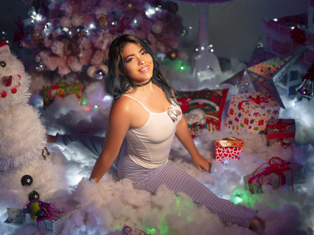KimLafayette tem seios de tamanho médio, veste uma camiseta branca e calças listradas de roxo, e está sentada em um "neve" branca e fofa, com uma árvore de Natal e presentes coloridos ao seu redor..