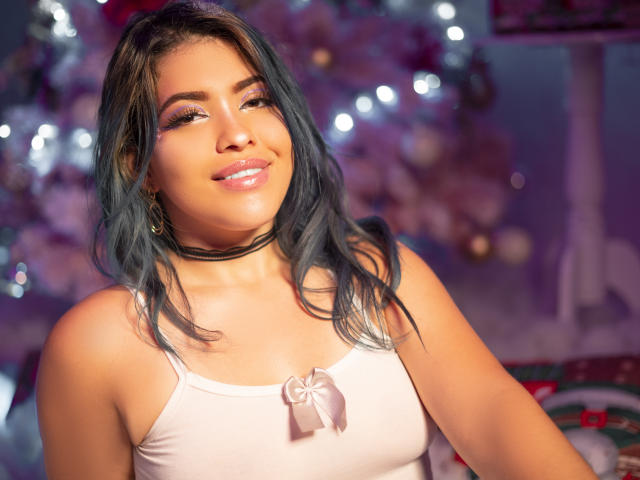 KimLafayette, con i capelli blu, sorridente, indossa una canotta bianca con un fiocco rosa, una collana nera e orecchini dorati, in un contesto natalizio con un albero decorato..