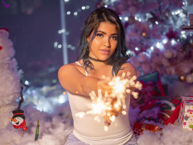KimLafayette with blue hair, white tank top, and choker, holding a sparkler, reaching forward, in a festive, snowy, and colorful setting with Christmas decorations.