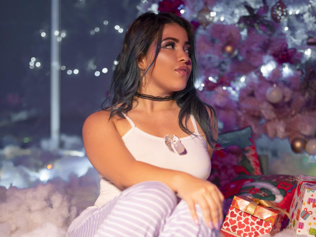 KimLafayette, com cabelo azul, vestindo uma camiseta branca e calças listradas, está sentada perto de uma árvore de Natal, olhando para o lado, com um presente na frente dela. Seios visíveis..