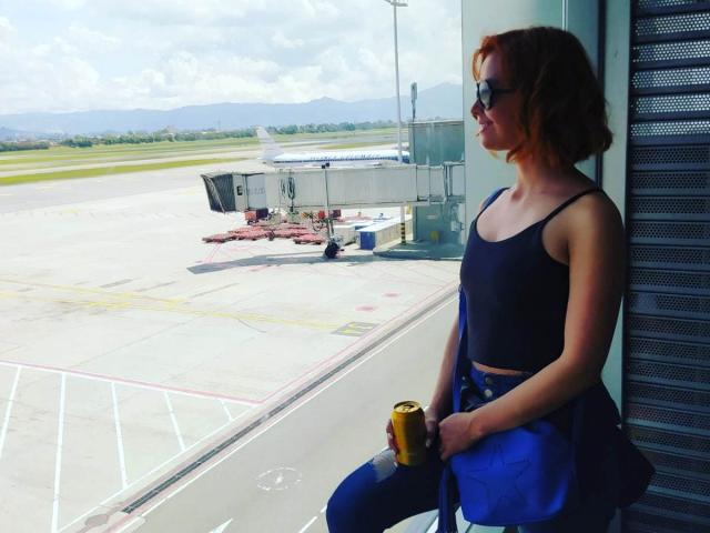 Eliza Jones, met rood haar, draagt een zwarte tanktop en blauwe jeans, ze houdt een blikje bier vast en staat bij een raam, kijkend naar buiten naar het vliegveld..