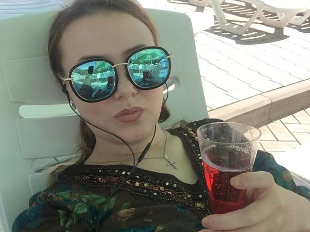 HanterHellen, avec des lunettes de soleil vertes réfléchissantes, des lèvres pincées et des écouteurs, tenant une boisson rouge dans un gobelet en plastique, portant un haut à motifs floraux, allongée sur une chaise blanche..