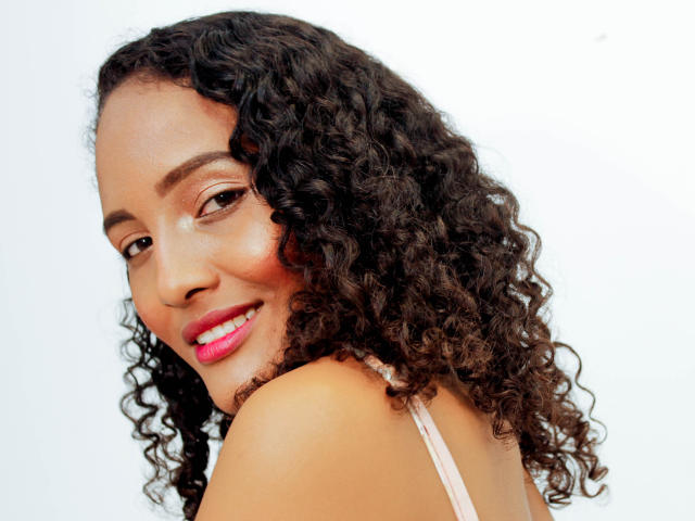 Megan Gil ha i capelli ricci e castano scuro, un sorriso carino con rossetto rosa, e sta guardando oltre la spalla. Indossa una spallina rosa, che mostra la sua spalla nuda..
