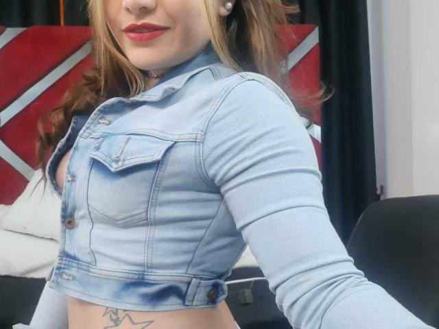 Sharlote Durant trägt eine hellblaue Jeansjacke im Crop-Stil, rote Lippenfarbe und hat ein Tattoo unter ihrer Brust. Ihr braunes Haar ist offen, und sie trägt Perlenohrringe..