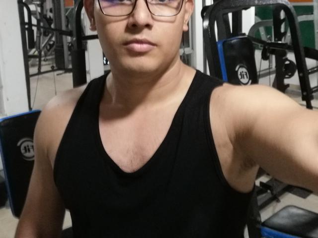 PatrickJean, met een bril, zwart tanktop, gespierde armen, lichtbruine huid, kort donker haar, licht gezwollen lippen, in een sportschool, die een selfie maakt..
