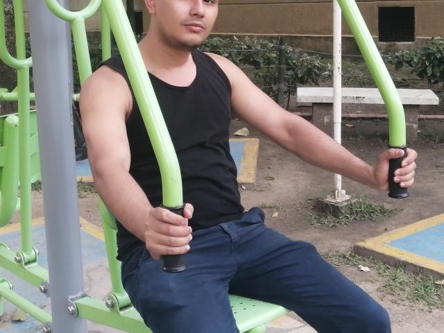 PatrickJean, muscular, tan-skinned, in black tank top and blue pants, gripping green metal bars of a playground, sitting with a relaxed, confident expression.