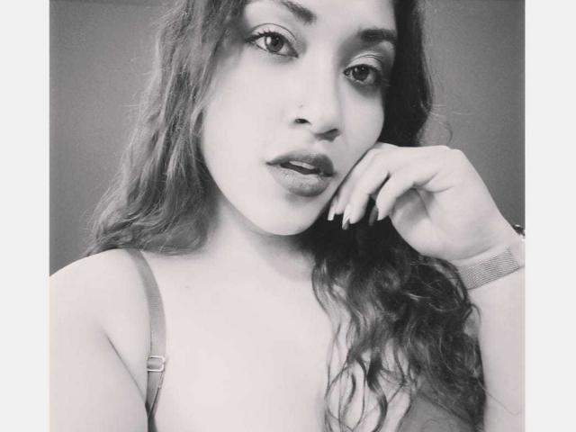 AmelyKent tiene el pelo ondulado, un piercing en la nariz y una expresión relajada, con la mano cerca de la boca, vistiendo un sujetador de tirantes y un reloj en la muñeca. Foto en blanco y negro..