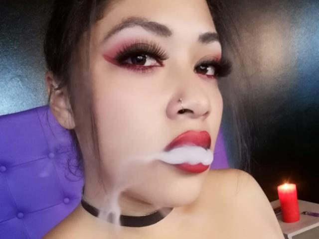 AmelyKent hat einen dicken roten Lidschatten, lange schwarze Wimpern und roten Lippenstift. Weiße Flüssigkeit befindet sich auf ihrer Unterlippe und ihrem Kinn. Sie trägt eine schwarze Halskette und einen Nasenring..