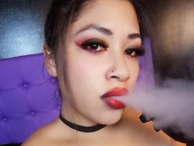 AmelyKent, con maquillaje rojo, un collarín negro y lápiz labial rojo, tiene un pequeño piercing en la nariz y parece estar fumando cerca de su boca. Tiene un reposacabezas morado, cabello oscuro y ojos ahumados..
