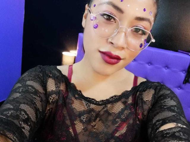 AmelyKent con maquillaje en forma de punto morado en la frente, usando gafas transparentes, una blusa de encaje negro, un sujetador rojo y lápiz labial oscuro, sentada en una cama morada con capitonado. Una vela en el fondo..