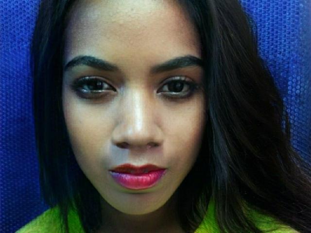 TafitaSexy has dark, straight hair, brown eyes, and shiny red lips. She's looking down with a slight smile, wearing a green top, against a blue textured background.