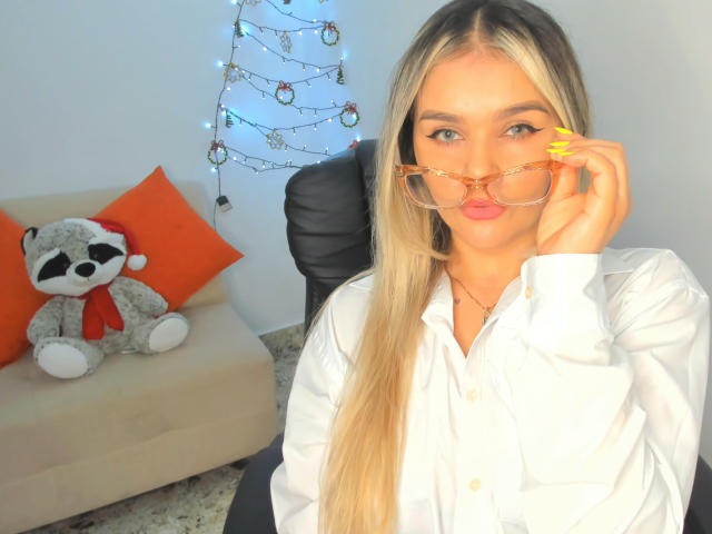 ValerieStone with long blonde hair, wearing a white shirt, holding orange glasses, yellow nails, and a cute ass, sitting next to a stuffed raccoon on a beige sofa.