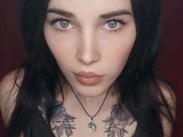 SonataStessi hat eine blasse Haut, graue Augen, schwarzes Haar und trägt ein schwarzes Oberteil mit Blattform-Tattoos unterhalb des Halses. Sie trägt eine Halskette mit einem Anhänger, hat volle Lippen und einen ernsten Gesichtsausdruck..