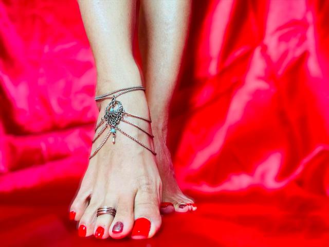 Die Füße von MistressCruelX sind mit rotem Nagellack lackiert, sie trägt eine silberne Fußkette und einen Ring am großen Zeh und steht auf einem leuchtend roten Stoff. Brüste, Hintern, Vulva usw. sind nicht sichtbar..