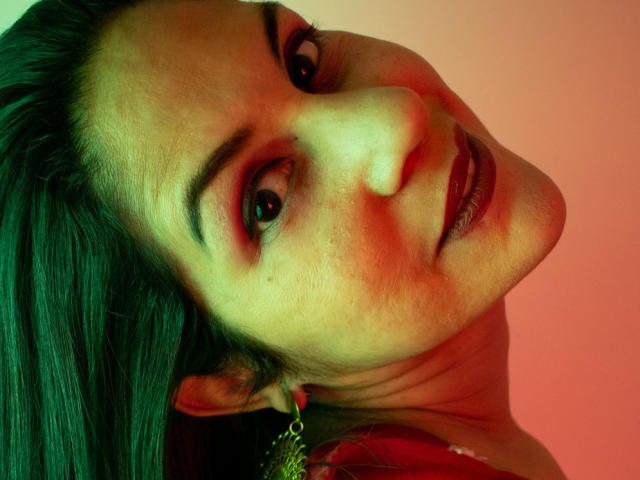 Le visage de RoraimAnaggie, avec ses cheveux verts, son éclairage rouge et vert, et un léger sourire. Elle a des taches de rousseur, des yeux foncés, et une boucle d'oreille dorée, et sa tête est légèrement inclinée vers la droite..