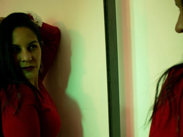 RoraimAnaggie with dark hair, red shirt, and hands behind head, looks in mirror. Her reflection shows her smiling, with green and yellow lighting. Her tits are visible.