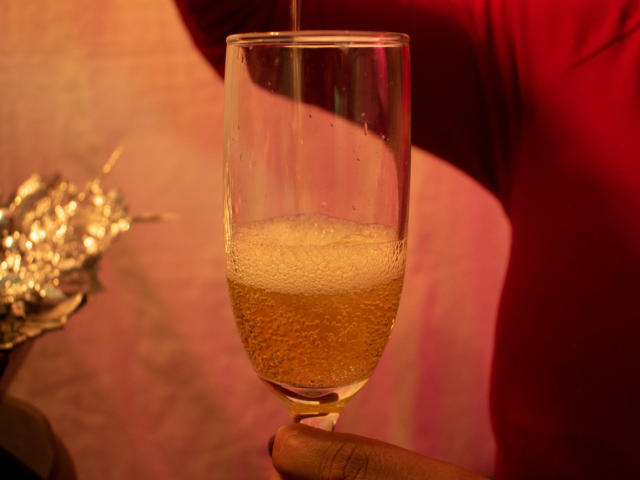 RoraimAnaggie pours bubbly champagne into a glass, showing a foamy, golden liquid with small bubbles. RoraimAnaggie's red sleeve is visible. Hand is holding the glass.