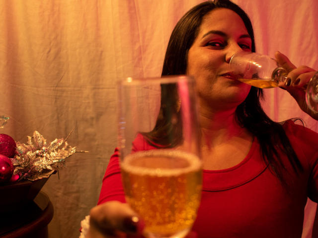 RoraimAnaggie with long black hair, red shirt, drinking from a glass of golden beer, holding another glass in foreground, red light, silver ornament, red ornament, slightly smiling.
