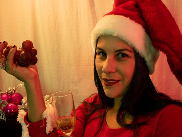RoraimAnaggie with dark hair, red Santa hat, and red shirt, holding grapes in one hand and a glass of white wine in the other, smiling. Christmas decorations in background.