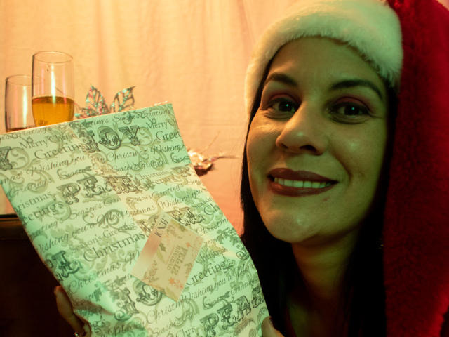 RoraimAnaggie with dark hair, wearing a red Santa hat, smiling, holding a festive paper, has a glass of orange drink on the left. She's got a cute, sexy face.